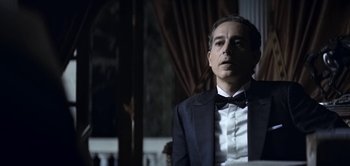 Movie still from “The Spy” (2019), directed by Gideon Raff – A man in a suit and bow tie sitting in front of a window; Medium shot, Low angle