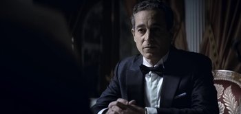 Movie still from “The Spy” (2019), directed by Gideon Raff – A man in a suit and bow tie sitting in a room; Medium shot, Over the shoulder angle