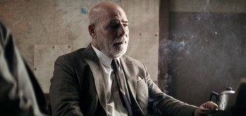 Movie still from “The Spy” (2019), directed by Gideon Raff – An older man wearing a suit and tie smoking a cigarette; Close Up shot, Low angle
