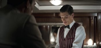 Movie still from “The Spy” (2019), directed by Gideon Raff – A man wearing a striped vest standing in front of another man; Medium shot, High angle