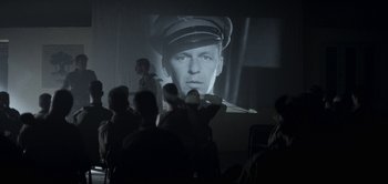 Movie still from “The Spy” (2019), directed by Gideon Raff – A group of people in a room watching a movie on a projector screen; Medium shot, Over the shoulder angle