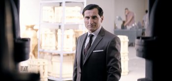 Movie still from “The Spy” (2019), directed by Gideon Raff – A man in a suit and tie standing in a room; Medium shot, Over the shoulder angle