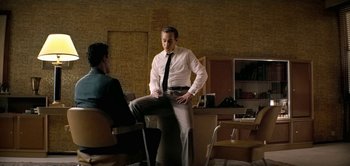 Movie still from “The Spy” (2019), directed by Gideon Raff – A man in a tie standing next to another man in a chair; Medium shot, Over the shoulder angle