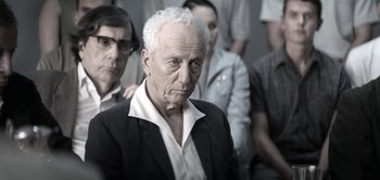Movie still from “The Spy” (2019), directed by Gideon Raff – An older man in a suit and tie with other men; Medium shot, Over the shoulder angle