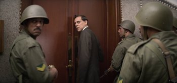 Movie still from “The Spy” (2019), directed by Gideon Raff – A man wearing a suit and tie standing in front of a door; Medium shot, Over the shoulder angle