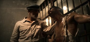 Movie still from “The Spy” (2019), directed by Gideon Raff – A man with a beard and a hat is standing next to a man with blood all over his body; Medium shot, Over the shoulder angle