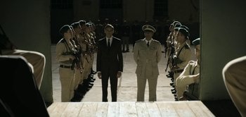Movie still from “The Spy” (2019), directed by Gideon Raff – Two men in suits standing next to each other in front of a group of soldiers; Wide shot, High angle