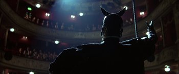 Movie still from “Batman Begins” (2005), directed by Christopher Nolan – A man with horns on his head is looking at a stage; Medium shot, Low angle