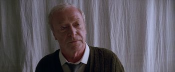 Movie still from “Batman Begins” (2005), directed by Christopher Nolan – An older man wearing a sweater and a tie; Close Up shot, Low angle