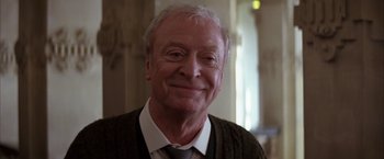 Movie still from “Batman Begins” (2005), directed by Christopher Nolan – An older man wearing a sweater and a tie; Close Up shot, Low angle
