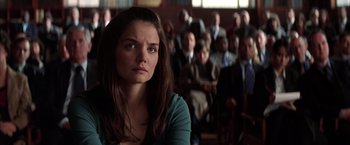 Movie still from “Batman Begins” (2005), directed by Christopher Nolan – A woman sitting in front of a group of people; Close Up shot, Over the shoulder angle