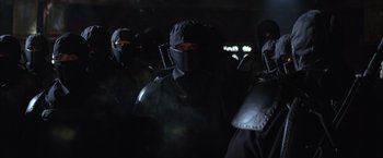 Movie still from “Batman Begins” (2005), directed by Christopher Nolan – A group of people in black suits and masks; Medium shot, Low angle