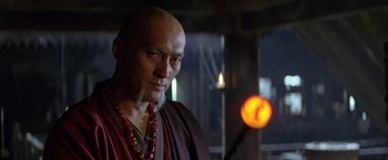 Movie still from “Batman Begins” (2005), directed by Christopher Nolan – A bald man wearing a red necklace and a red robe; Close Up shot, Low angle