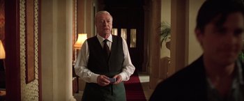 Movie still from “Batman Begins” (2005), directed by Christopher Nolan – An older man standing in front of a red carpet; Medium shot, Over the shoulder angle