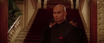 Movie still from “Batman Begins” (2005), directed by Christopher Nolan – A bald man in a black suit standing in front of a staircase; Close Up shot, Low angle