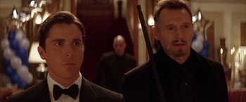 Movie still from “Batman Begins” (2005), directed by Christopher Nolan – A man in a suit and tie holding a cane in front of another man in a suit and tie; Close Up shot, Low angle