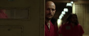 Movie still from “Batman Begins” (2005), directed by Christopher Nolan – A bald man in a red shirt is looking at the camera; Close Up shot, Over the shoulder angle