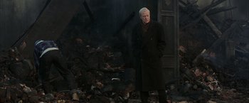 Movie still from “Batman Begins” (2005), directed by Christopher Nolan – An older man standing in front of a pile of rubble; Medium shot, High angle