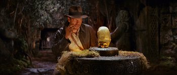 Movie still from “Raiders of the Lost Ark” (1981), directed by Steven Spielberg – A man sitting in front of an egg on top of a table; Medium shot, Low angle