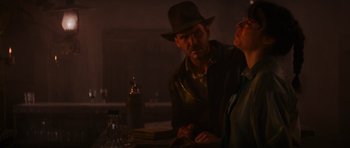 Movie still from “Raiders of the Lost Ark” (1981), directed by Steven Spielberg – A man and a woman sitting at a table in front of a bottle; Medium shot, Over the shoulder angle