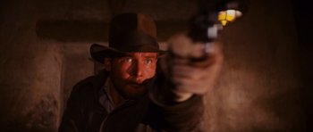 Movie still from “Raiders of the Lost Ark” (1981), directed by Steven Spielberg – A man in a hat and jacket holding a gun; Close Up shot, Over the shoulder angle