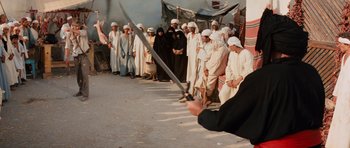 Movie still from “Raiders of the Lost Ark” (1981), directed by Steven Spielberg – A group of people standing in front of a man holding a sword; Wide shot, Over the shoulder angle