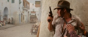 Movie still from “Raiders of the Lost Ark” (1981), directed by Steven Spielberg – A man holding a gun while standing next to a wall; Medium shot, Over the shoulder angle
