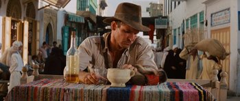 Movie still from “Raiders of the Lost Ark” (1981), directed by Steven Spielberg – A man sitting at a table drinking from a cup; Medium shot, Low angle