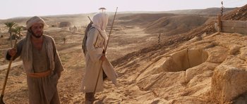 Movie still from “Raiders of the Lost Ark” (1981), directed by Steven Spielberg – A man in a long white coat walking through the desert; Wide shot, Low angle