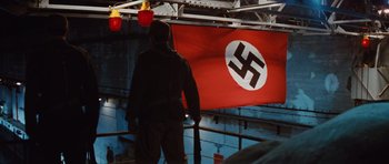 Movie still from “Raiders of the Lost Ark” (1981), directed by Steven Spielberg – A man holding a rifle in front of a nazi flag; Wide shot, Low angle