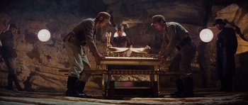 Movie still from “Raiders of the Lost Ark” (1981), directed by Steven Spielberg – A group of men standing next to a table in a cave; Wide shot, High angle