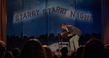 Movie still from “The Squid and the Whale” (2005), directed by Noah Baumbach – A man standing on a chair in front of an audience; Wide shot, High angle
