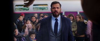 Movie still from “The Takedown” (2022), directed by Louis Leterrier – A man in a suit and tie standing in front of a group of people; Close Up shot, Over the shoulder angle