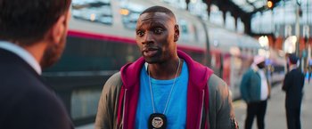 Movie still from “The Takedown” (2022), directed by Louis Leterrier – A man with a beard and a blue t - shirt is standing in front of a train; Close Up shot, Over the shoulder angle