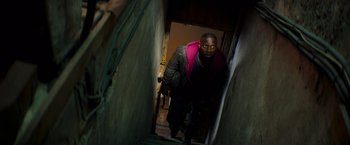 Movie still from “The Takedown” (2022), directed by Louis Leterrier – A man in a pink vest is walking down the stairs; Medium shot, High angle