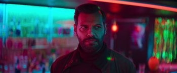 Movie still from “The Takedown” (2022), directed by Louis Leterrier – A man with a beard is standing in a room with neon lights in the background; Close Up shot, Low angle
