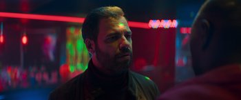 Movie still from “The Takedown” (2022), directed by Louis Leterrier – A man with a beard is standing in a dark room; Close Up shot, Over the shoulder angle