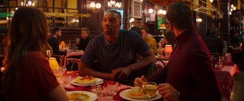 Movie still from “The Takedown” (2022), directed by Louis Leterrier – A man sitting at a table with a plate of food in front of him; Medium shot, Over the shoulder angle