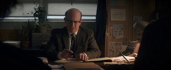 Movie still from “The Trial of the Chicago 7” (2020), directed by Aaron Sorkin – An older man sitting at a desk writing on a piece of paper; Medium shot, Over the shoulder angle