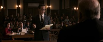 Movie still from “The Trial of the Chicago 7” (2020), directed by Aaron Sorkin – A man in a suit and tie holding a sheet of paper in front of an audience; Medium shot, Over the shoulder angle