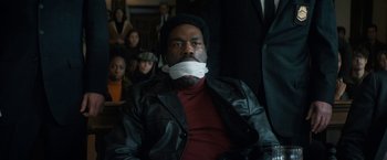 Movie still from “The Trial of the Chicago 7” (2020), directed by Aaron Sorkin – A man with his mouth taped shut sitting in front of a group of people; Medium shot, High angle