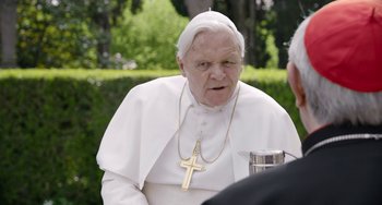 Movie still from “The Two Popes” (2019), directed by Fernando Meirelles – An older man wearing a white outfit and a cross necklace; Close Up shot, Over the shoulder angle