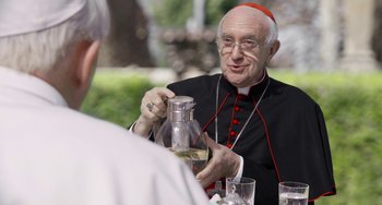 Movie still from “The Two Popes” (2019), directed by Fernando Meirelles – An older man wearing glasses and a red and white hat; Close Up shot, Over the shoulder angle
