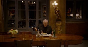 Movie still from “The Two Popes” (2019), directed by Fernando Meirelles – An older man sitting at a table with a plate in front of him; Wide shot, High angle