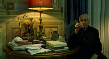 Movie still from “The Two Popes” (2019), directed by Fernando Meirelles – An older man sitting at a desk with papers and a clock; Medium shot, High angle
