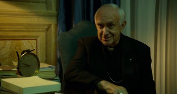 Movie still from “The Two Popes” (2019), directed by Fernando Meirelles – An older man sitting at a desk in a chair; Close Up shot, High angle