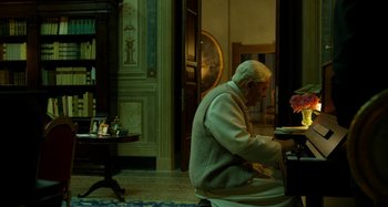 Movie still from “The Two Popes” (2019), directed by Fernando Meirelles – An older man sitting in a room with a book; Wide shot, High angle