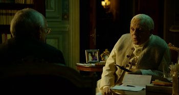Movie still from “The Two Popes” (2019), directed by Fernando Meirelles – An older man sitting at a table in front of another man; Medium shot, Over the shoulder angle