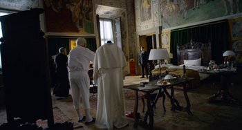 Movie still from “The Two Popes” (2019), directed by Fernando Meirelles – A man in a white suit stands next to a woman in a white suit; Wide shot, Over the shoulder angle