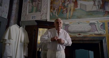 Movie still from “The Two Popes” (2019), directed by Fernando Meirelles – An older man in a white shirt is standing in front of a mural; Medium shot, Over the shoulder angle